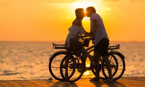 couple with bicycles kiss on boardwalk in front of the setting sun